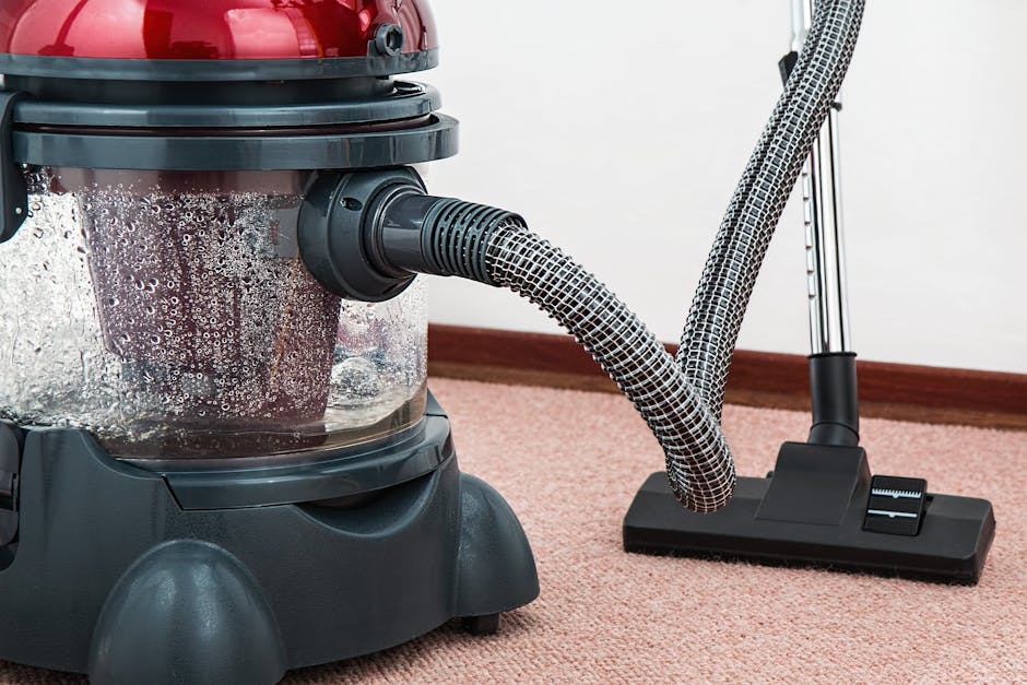 A woman with curly hair, dressed in a beige jacket and jeans, is performing surface cleaning on a patterned area rug in a living room, using a yellow vacuum cleaner. She is bent slightly forward, hold